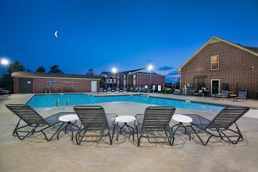 A pool with chairs and a building in the background at night.