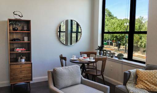 A living room with a grey couch, a wooden bookshelf, a round mirror, and a window with a view of trees.