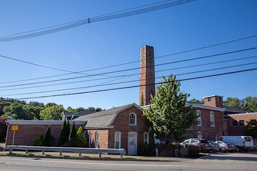 A brick building with a tall chimney is surrounded by trees and parked cars.