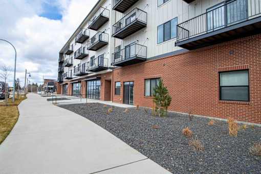 A sidewalk leads to a building with balconies and a small tree in front.