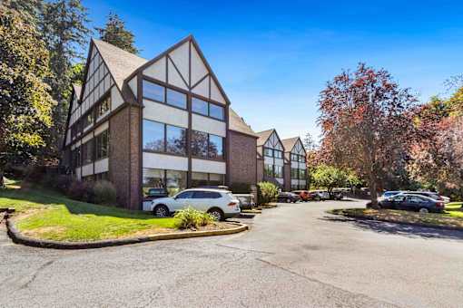 A modern building with a mix of brick and glass windows is surrounded by trees and has a parking lot in front.