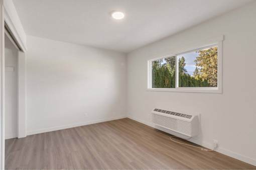 A room with a window showing trees and a white air conditioner on the floor.