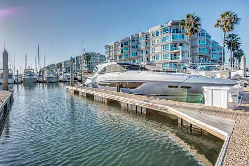 A yacht is docked at a marina with a building and palm trees in the background.