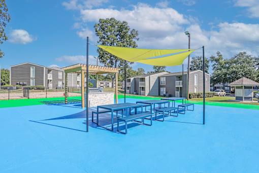 A picnic area with a blue surface and a yellow shade structure by the bbq grills at Maplewood apartments in Shreveport, LA.