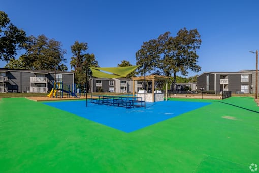 BBQ and picnic area with a blue and green surface and a yellow sunshade at Maplewood apartments in Shreveport, LA.