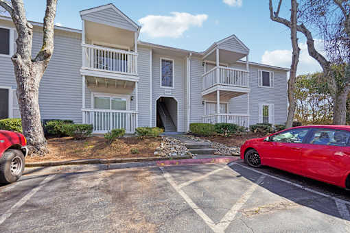 A red car is parked in a parking lot in front of a two-story apartment building.