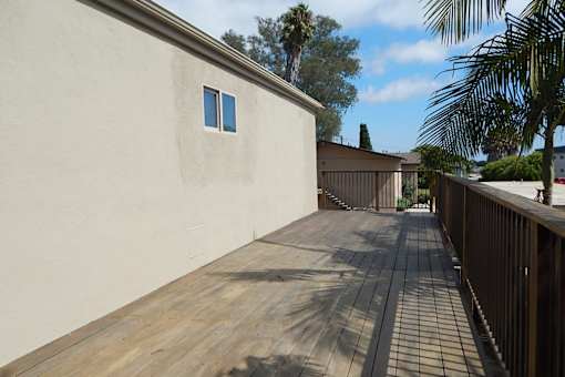 A wooden deck leads to a house with a white wall and a window.