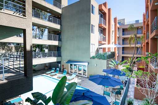 A pool area with blue umbrellas and chairs is surrounded by apartment buildings.