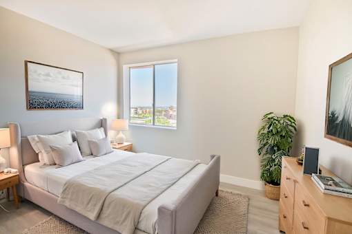 A bedroom with a large bed, a nightstand, a lamp, a plant, and a window.