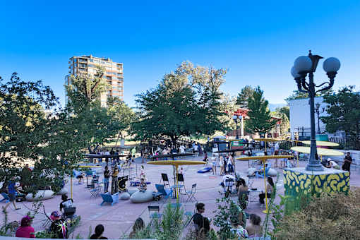 A park with people sitting and standing on the grass and a building in the background.