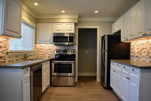 View of the kitchen, featuring wood like floors, stainless appliances, tile backsplash, white cabinets, window view over sink with wood-like blinds