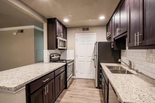 View into kitchen with stone counters, tile back splash, stainless appliances, and brown cabinets