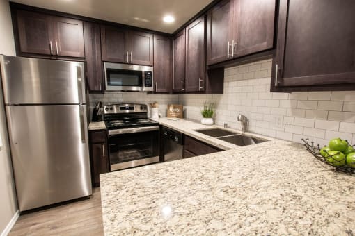 View of kitchen with stone counters, inlaid sink, stainless appliances, tile backsplash, brown cabinets, and wood look flooring.