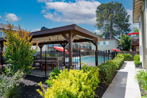 View into gated community pool with covered area that has a picnic table and seating areas, surrounded by lush landscaping