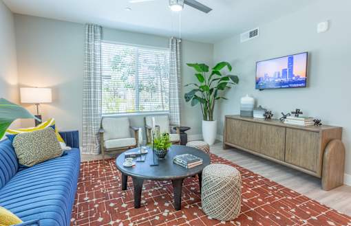 A living room with a blue couch, a coffee table, a TV, and a large window with a view of the city.