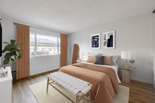 Goleta, CA, Apartments Near Target - Pacific Oaks - Bedroom with Wood-Style Flooring, a Bed, an Ottoman, Two Bedside Tables with Lamps, and a Large Window with Curtains.