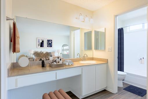 Goleta, CA, Apartments Near UCSB - Pacific Oaks - Bathroom with Wood-Style Flooring, a Spacious Granite Countertop Sink with White Cabinetry, a Large Mirror, and a Shower with a Tub and a Shower Curtain.