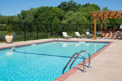 Outdoor pool surrounded by pool chairs and a fence, trees is the background