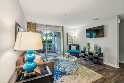 A blue lamp sits on a table in a room with a glass table and a sofa. at The Timbers Apartments, Richmond, VA