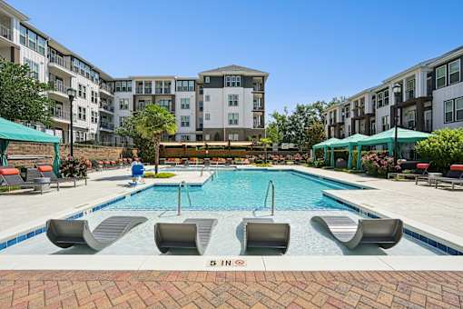 A swimming pool surrounded by lounge chairs and umbrellas in front of apartment buildings.