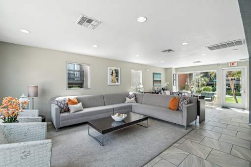 A grey couch with orange pillows sits in a living room with a glass coffee table in front of it.
