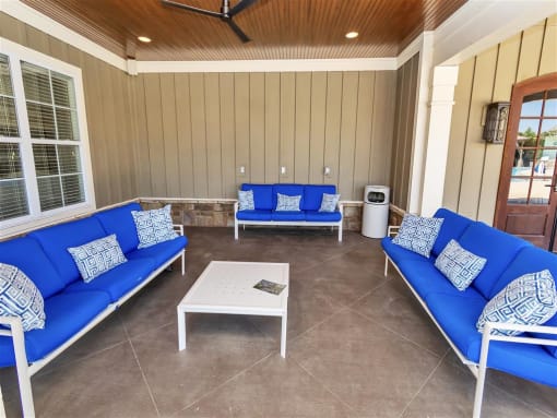 Three cushioned benches and a small table under a ceiling fan on the outdoor covered clubhouse patio