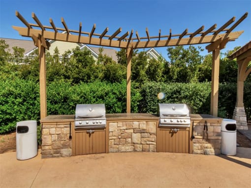 A large stone grilling station with two outdoor grills under a wooden pergola near trees
