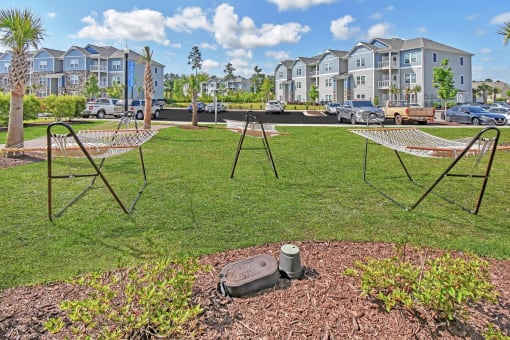 a swing set and two hammocks in a grassy area with apartment buildings in the background