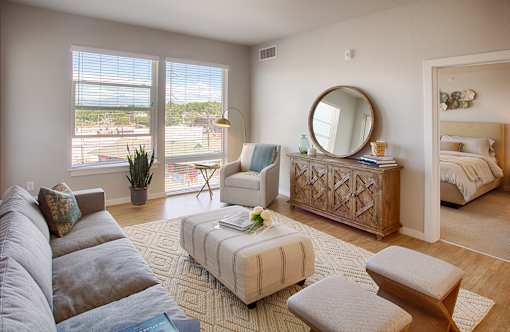 A well-lit, spacious living room with a grey couch, a white armchair, a wooden coffee table, and a large window with a view of the outside.
