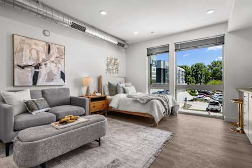 A bedroom with a bed, a couch, a nightstand, and a window overlooking a parking lot.