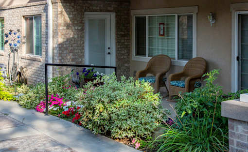 Two chairs are on a porch with a flower bed in front.