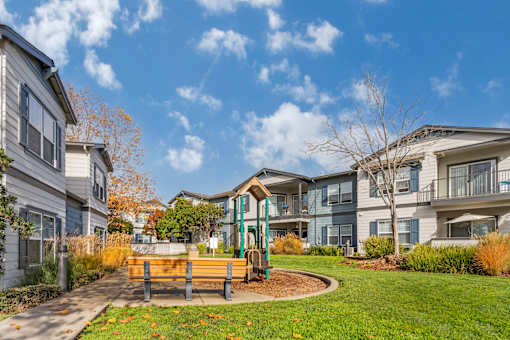 A playground with a slide and a bench is surrounded by a grassy area and apartment buildings.