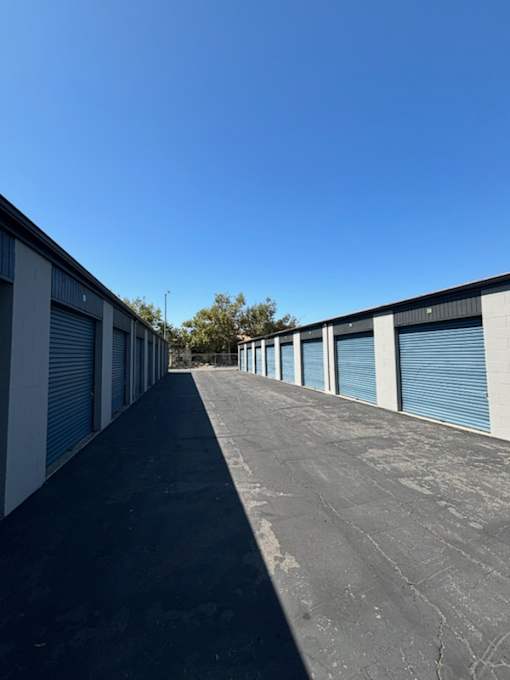 Outdoor storage facility with multiple units featuring blue roll-up doors and an asphalt driveway under a clear blue sky.