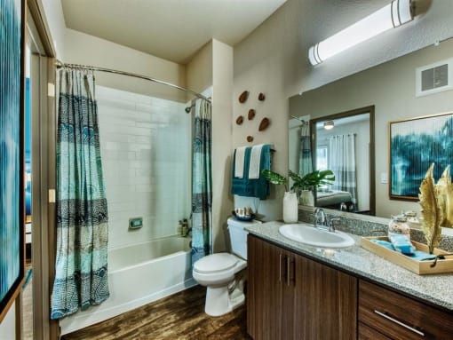 Apartment guest bathroom with white tub and blue shower curtain. Toilet can be seen next to a modern wood vanity and large mirror.at Craft at Gilbert and Baseline, Mesa  