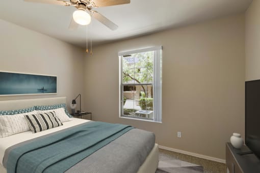 Photo of a bedroom with queen size bed and light gray & blue bed spread.  A window showing a sunny day can be seen in the background.