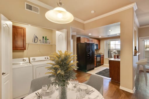 Photo of the kitchen from the dining area. The round dining table with 2 chairs can be seen. A bouquet of yellow flowers in a vase  sits on the dining table. A washer and dryer can be seen in a closet to the left with a folding door open showing the appliances can be hidden from view. A gloss black fridge and black stove can be seen in the kitchen.