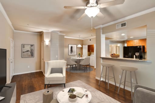 Photo of a model apartment living room and kitchen bar stool area with dining area in the background. The coffee table and white chair can be seen in the living room with bar stools facing the kitchen. A round dining table with four chairs is in the dining room.