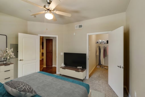 Photo of a master bedroom showing part of a queen size bed with gray bed spread. A flat screen TV sits on a wall facing the bed with small cabinet below the TV.  An open door to the right shows the walk in closet