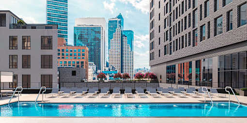 A swimming pool with sun loungers in the foreground and a city skyline in the background.