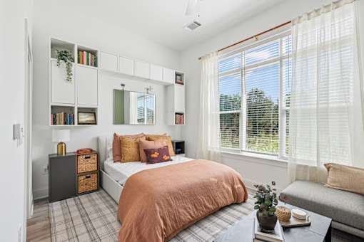 A bedroom with a bed, a window with blinds, a nightstand, a bookshelf, and a plant on a table at Link Apartments® CYKEL Apartments, Charlotte, NC, 28208