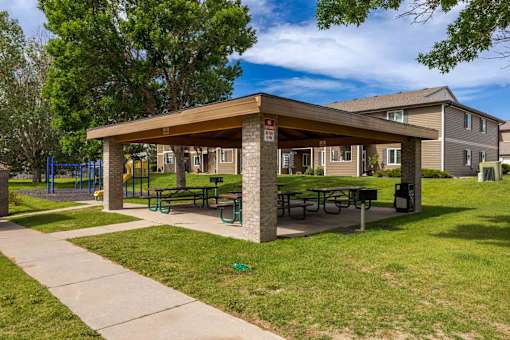 A covered picnic area with tables and benches is surrounded by green grass and trees.