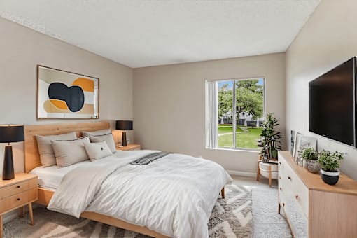 A bedroom with a large bed, a nightstand, and a window overlooking a green lawn.