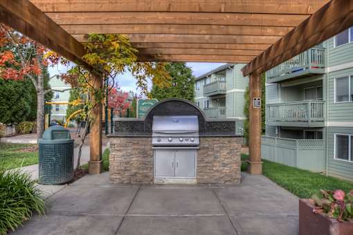 A stone and tile outdoor kitchen with a grill and sink under a wooden pergola.