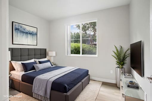 A bedroom with a bed, a nightstand, a lamp, a plant, a television, and a painting on the wall.