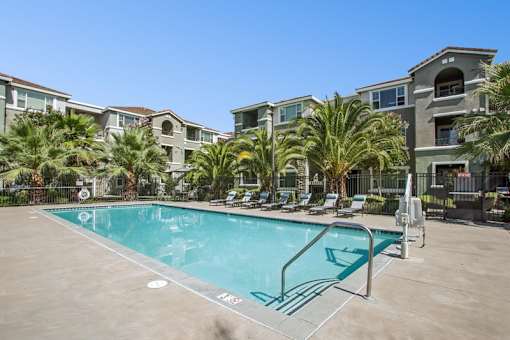 A swimming pool surrounded by palm trees and lounge chairs in front of apartment buildings.
