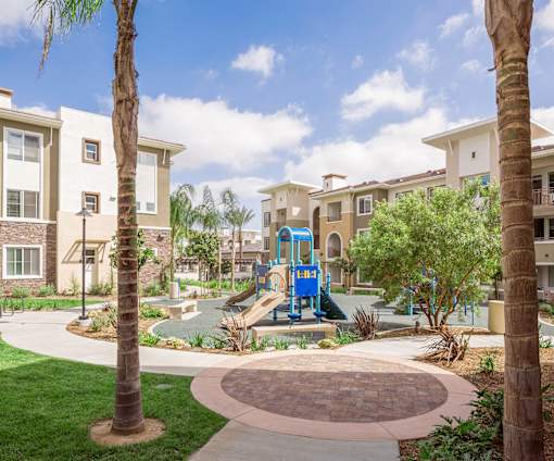 A playground with a blue slide is surrounded by palm trees and apartment buildings.