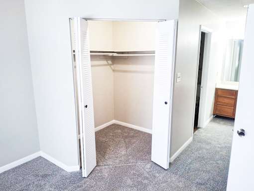 A white open closet with a carpeted floor and a wooden dresser in the hallway.