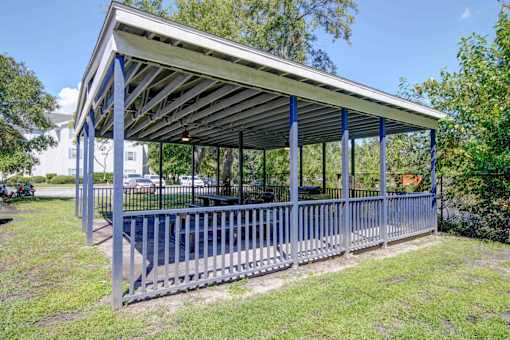 A pavilion with a metal roof and blue pillars is surrounded by a metal fence and grass.