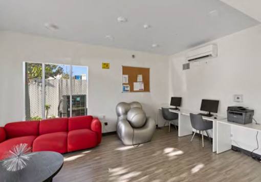 A red sofa and a grey chair are in a room with a white desk and a window.