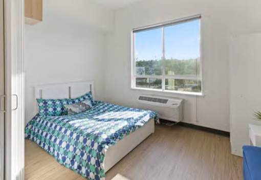 A bedroom with a bed, a window with a view of trees and a building, and a blue chair.